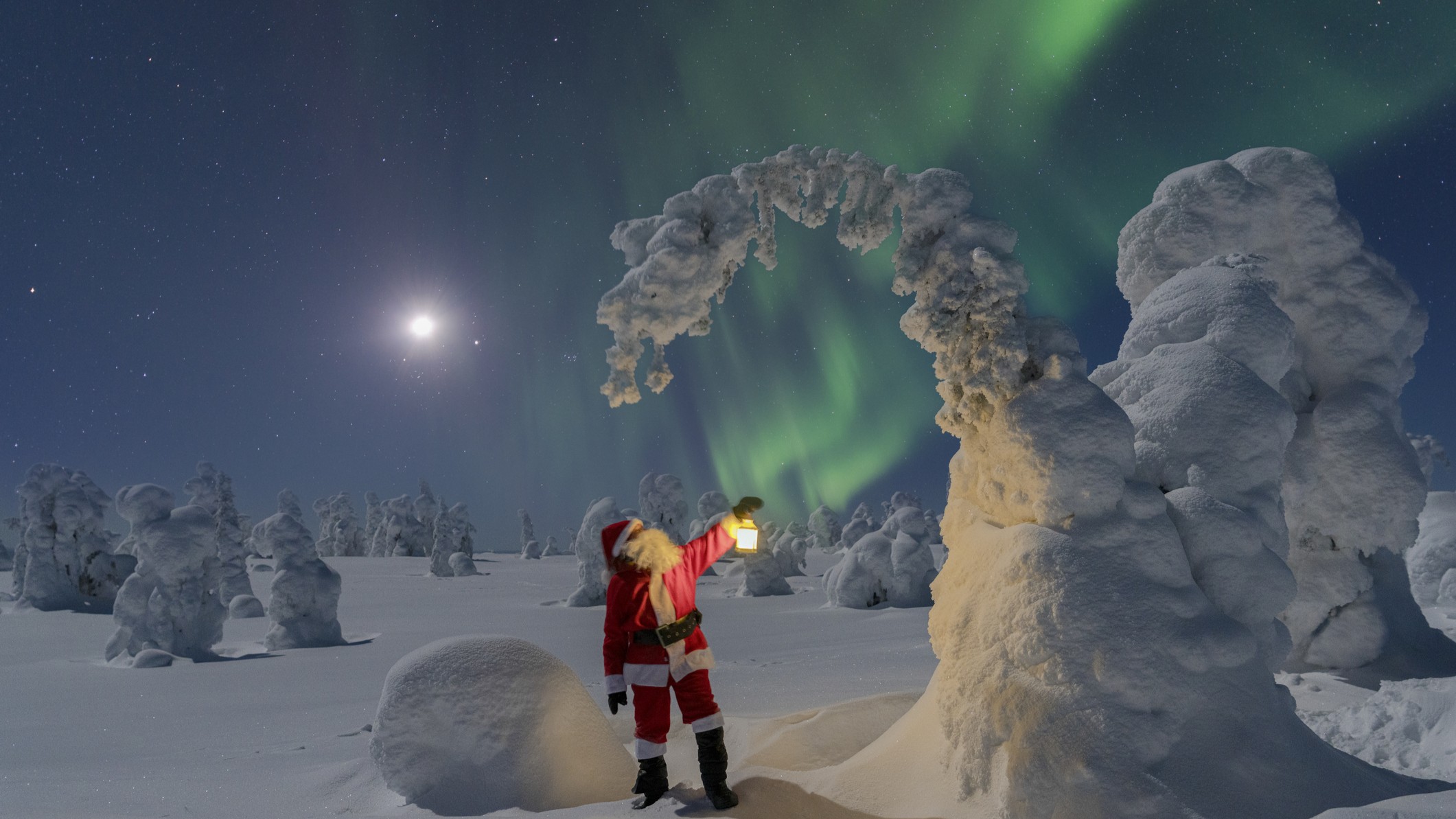 Santa Claus stands admiring snow swept trees as the northern lights dance behind as green curtains of light.