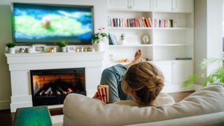 A woman is watching television in a living room, the television is placed above a fire place and she is eating popcorn.
