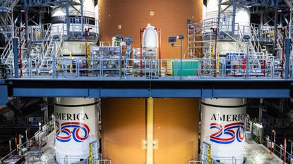 An image from inside a warehouse shows two tall white cylinders on either side of a yellow taller cylinder. The two white cylinders have red, white and blue paint with the words 'America 250' on them.