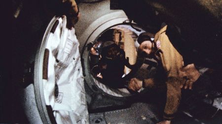 two men floating in space reach out and shake hands across the open threshold of a spacecraft hatchway